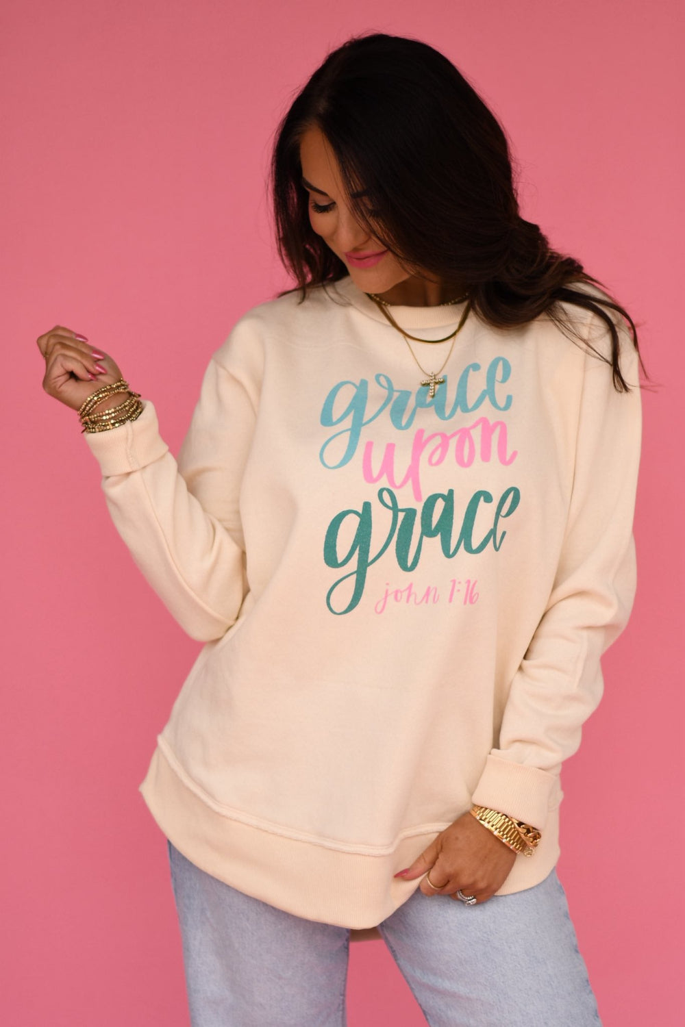 Woman wearing a cream-colored “Grace Upon Grace” Christian graphic tee with teal and pink lettering, styled with denim jeans against a pink background.