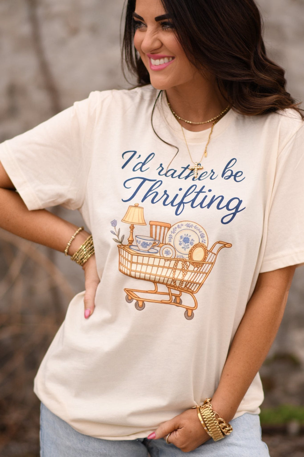 Woman wearing a natural colored graphic t-shirt that says “I’d Rather Be Thrifting” with a vintage shopping cart illustration, styled with jeans and gold jewelry