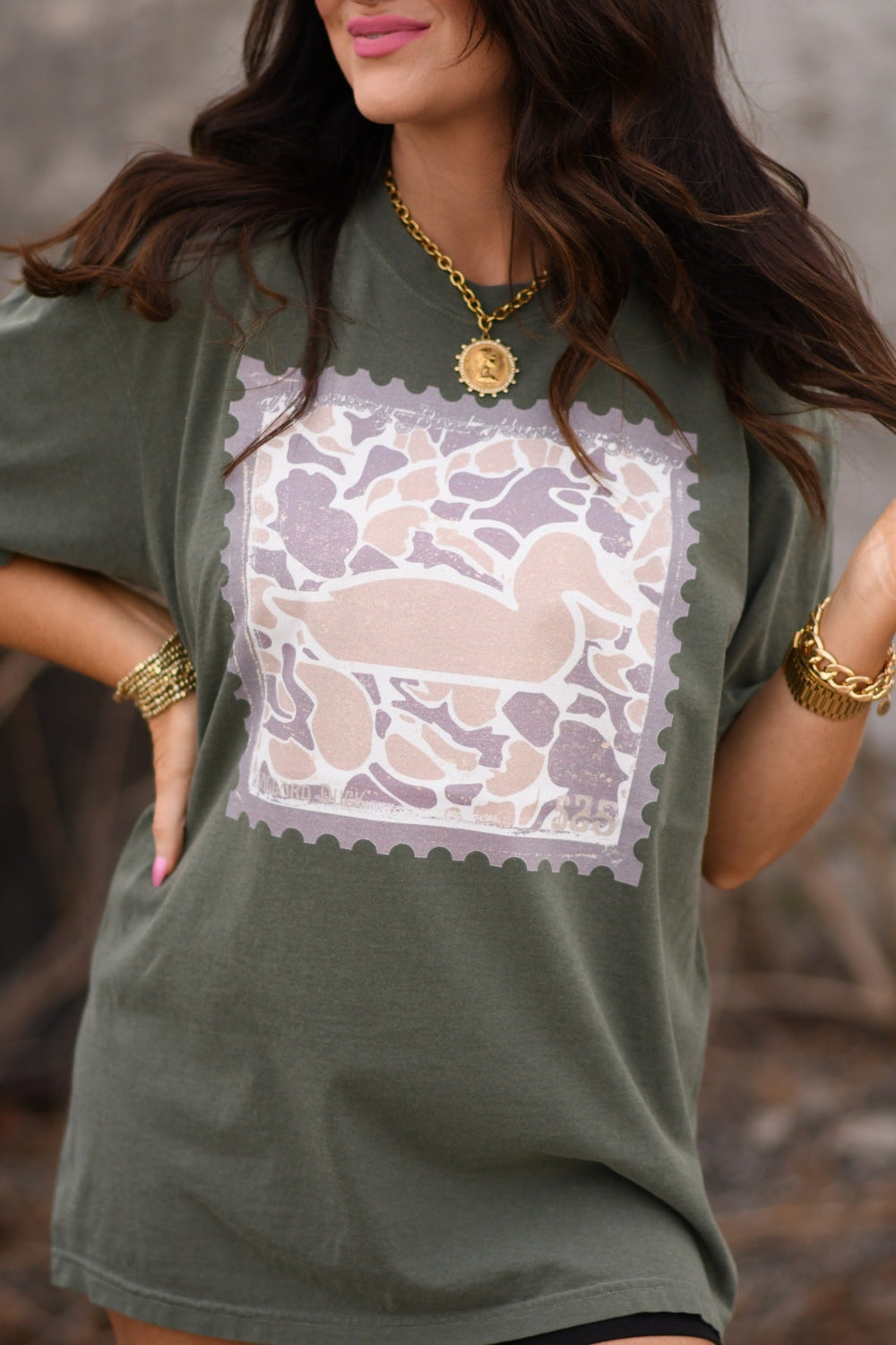 Woman wearing moss green Comfort Colors unisex t-shirt featuring duck camo stamp graphic, styled with sunglasses and gold jewelry.