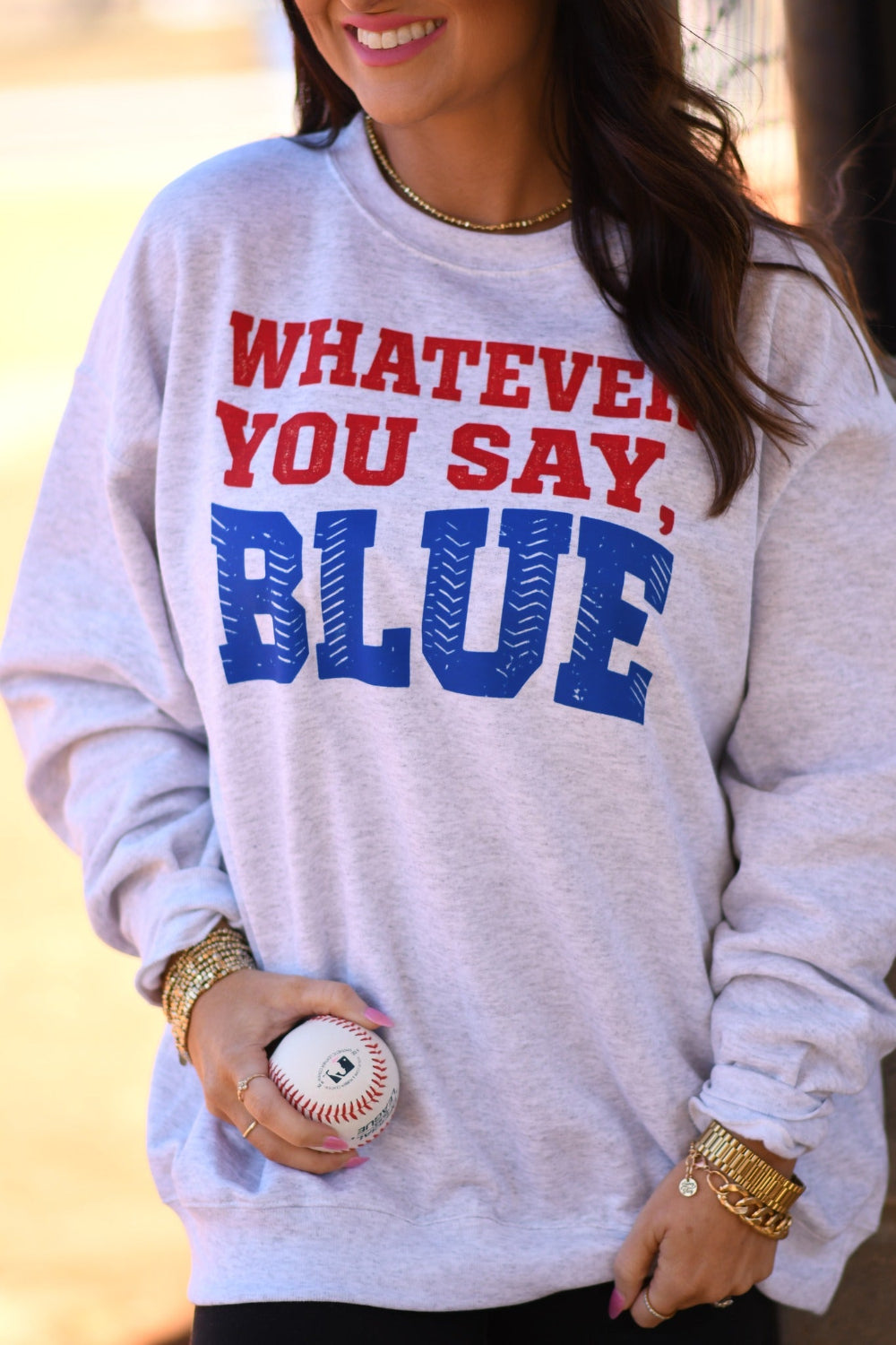 Woman wearing ash gray โWhatever You Say Blueโ baseball graphic sweatshirt with red and blue lettering, styled with a baseball cap and holding a baseball at a ball field.