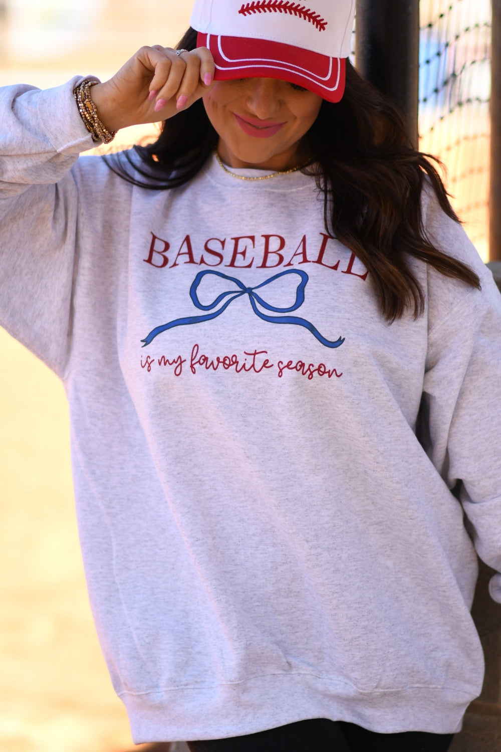 Woman wearing ash gray “Baseball Is My Favorite Season” graphic sweatshirt with blue bow design and red baseball hat at baseball field.