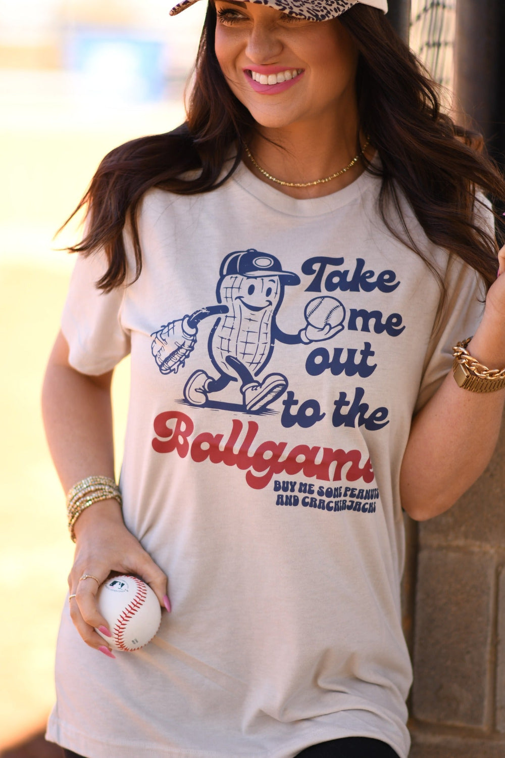 Woman wearing Heather Dust Bella Canvas βTake Me Out To The Ballgameβ peanut baseball graphic tee with leopard brim baseball hat at a baseball field.