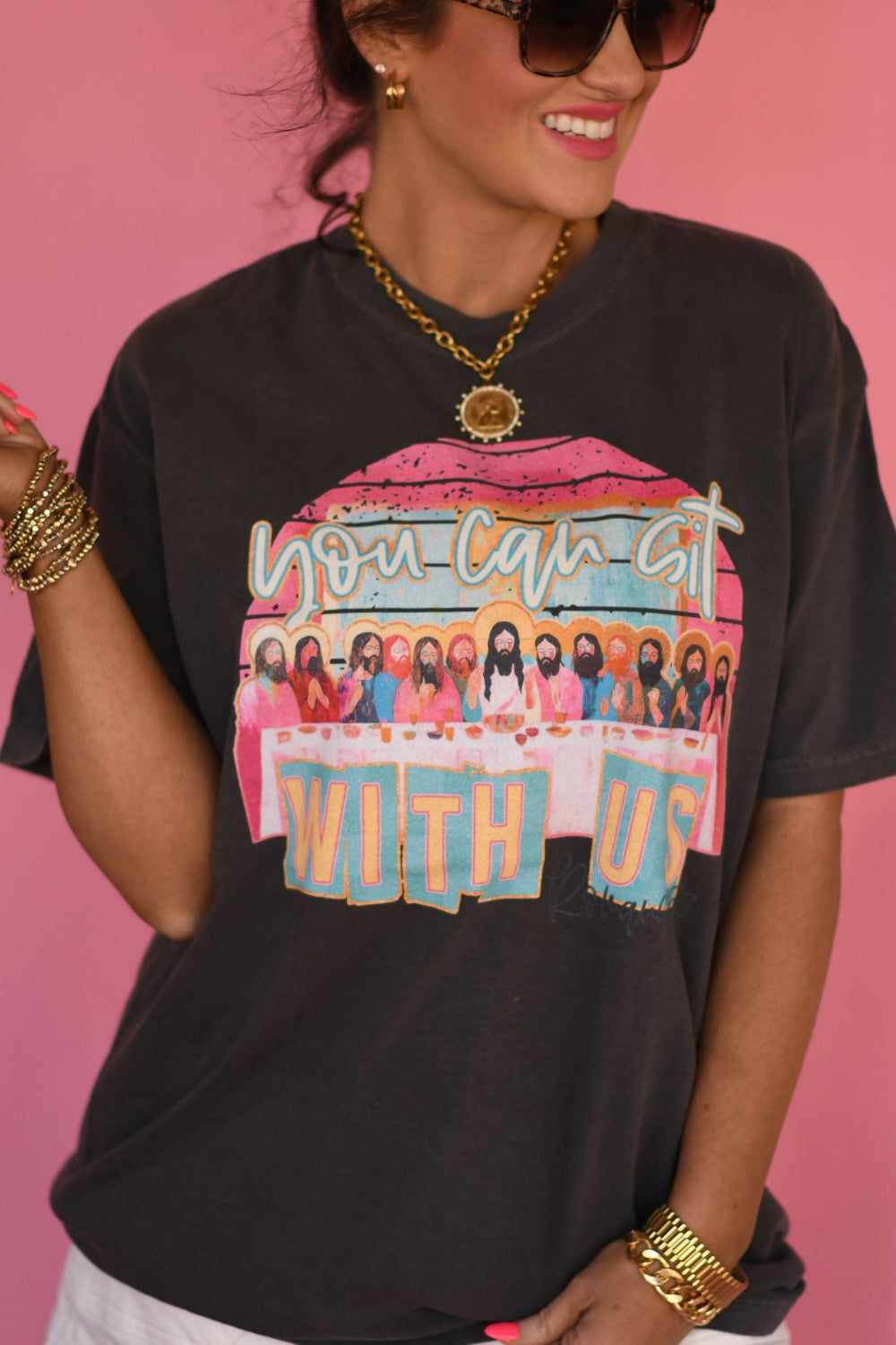 Woman wearing a Pepper Comfort Colors βYou Can Sit With Usβ Christian graphic tee with faith-based design, styled with white shorts against a pink background.