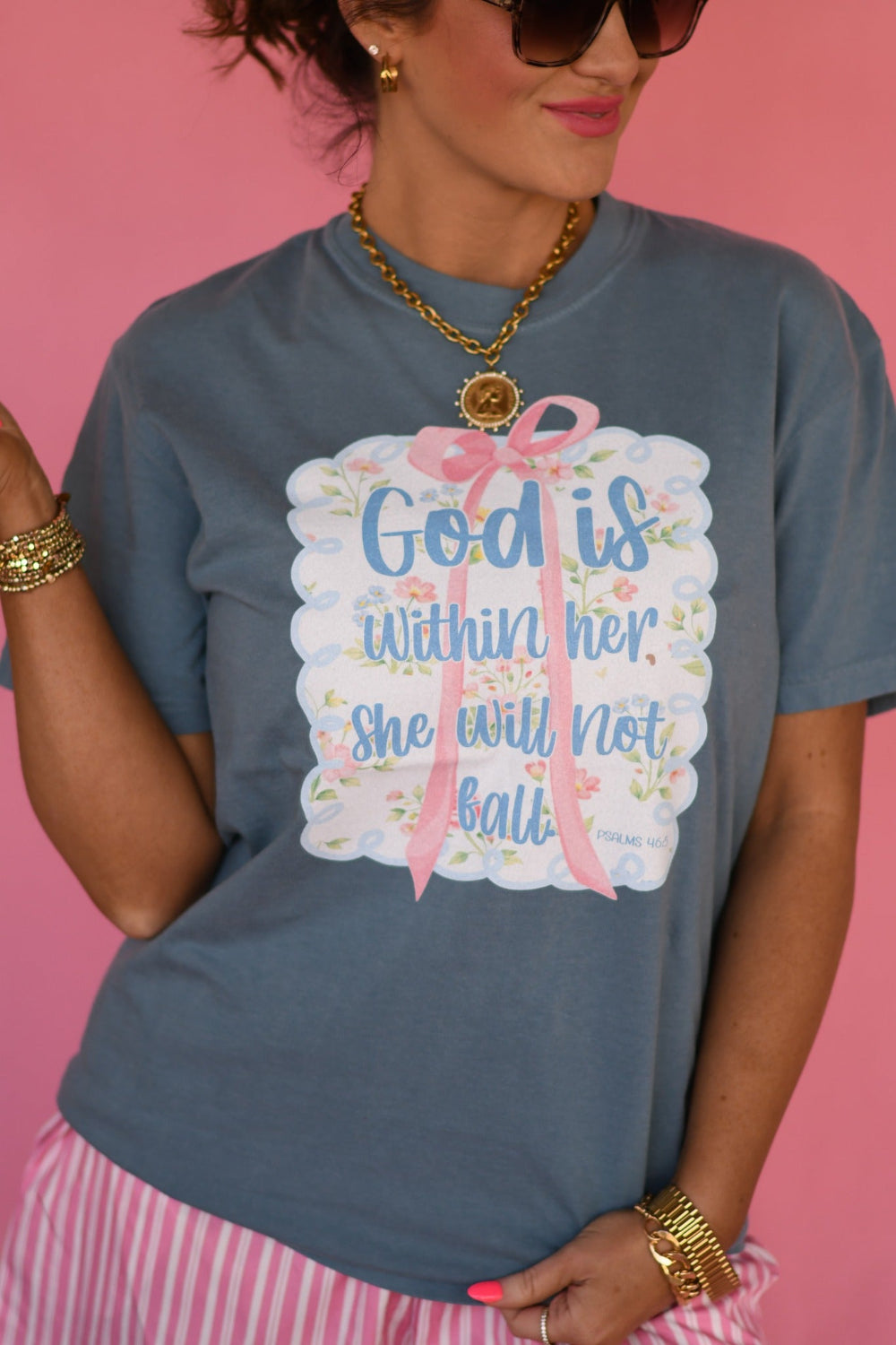 Woman wearing Ice Blue Comfort Colors βGod Is Within Her She Will Not Fallβ Christian graphic tee with pink bow floral design from Psalm 46:5