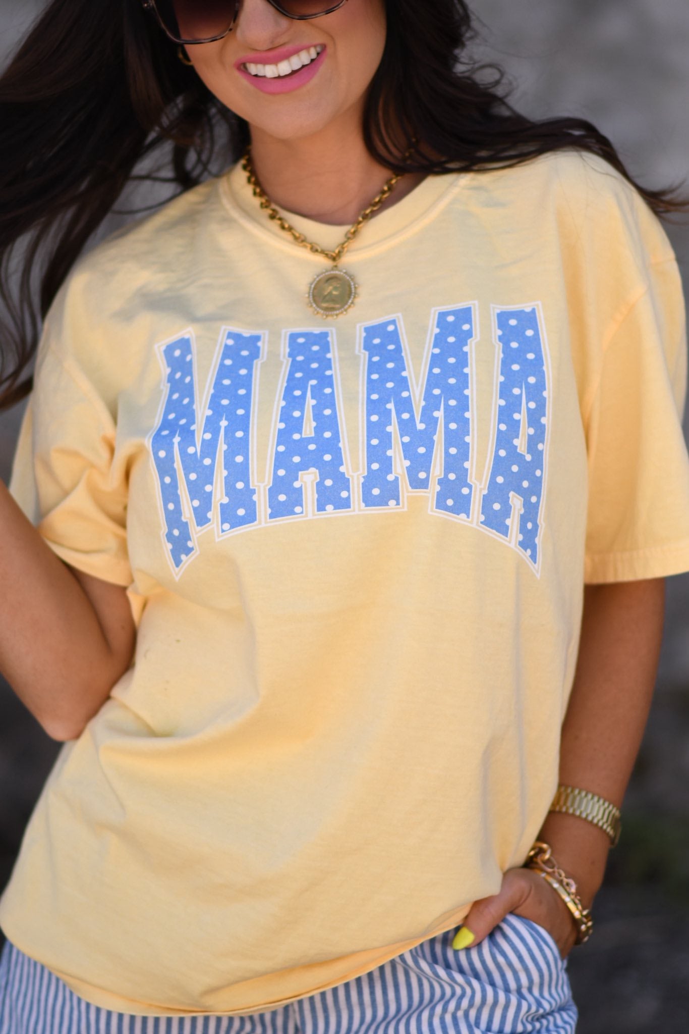 Woman wearing butter yellow MAMA graphic tee with blue polka dots paired with striped shorts and sunglasses