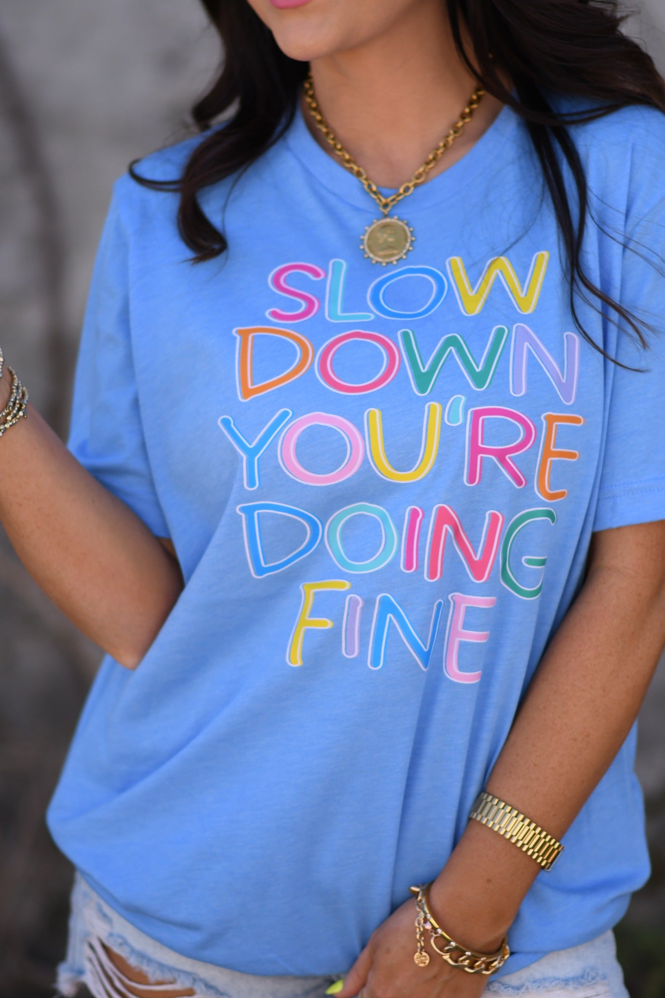 Woman wearing a heather Columbia blue graphic tee with colorful “Slow Down You’re Doing Fine” text paired with distressed denim shorts and sunglasses.