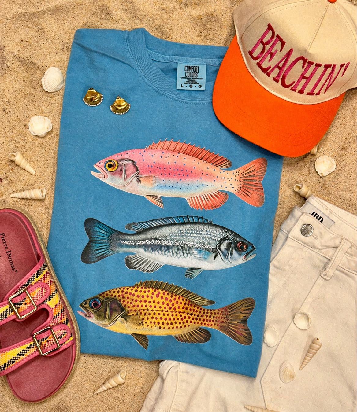 Royal Caribbean blue Comfort Colors fish graphic tee styled on sand with beach hat, white shorts, pink sandals, and seashells.