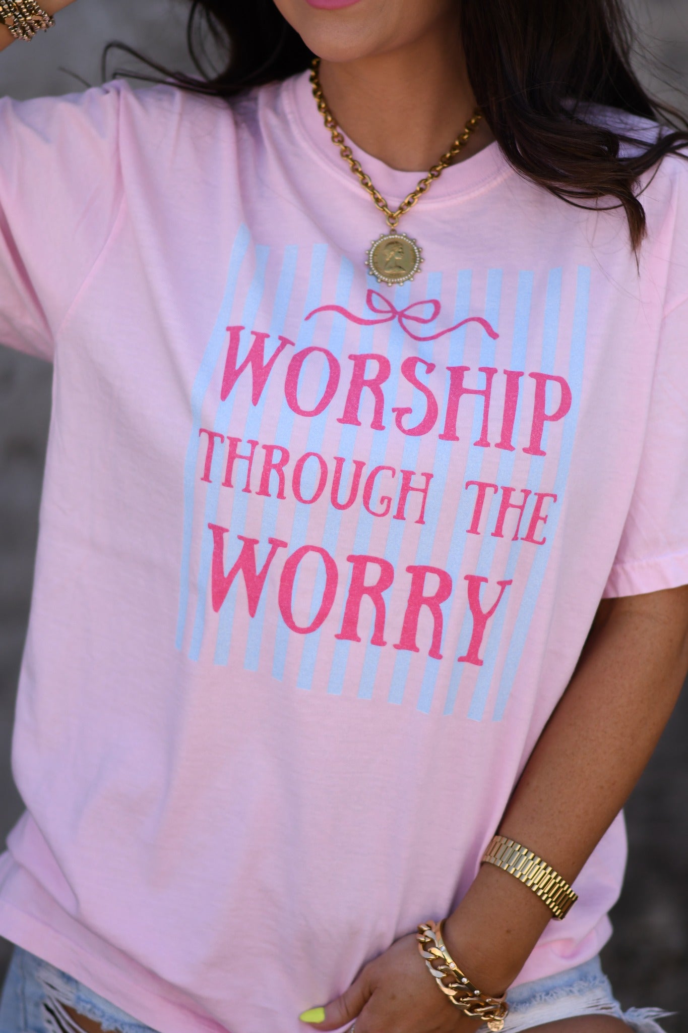 Woman wearing pink Worship Through The Worry Christian graphic tee with striped bow design and denim shorts outdoors