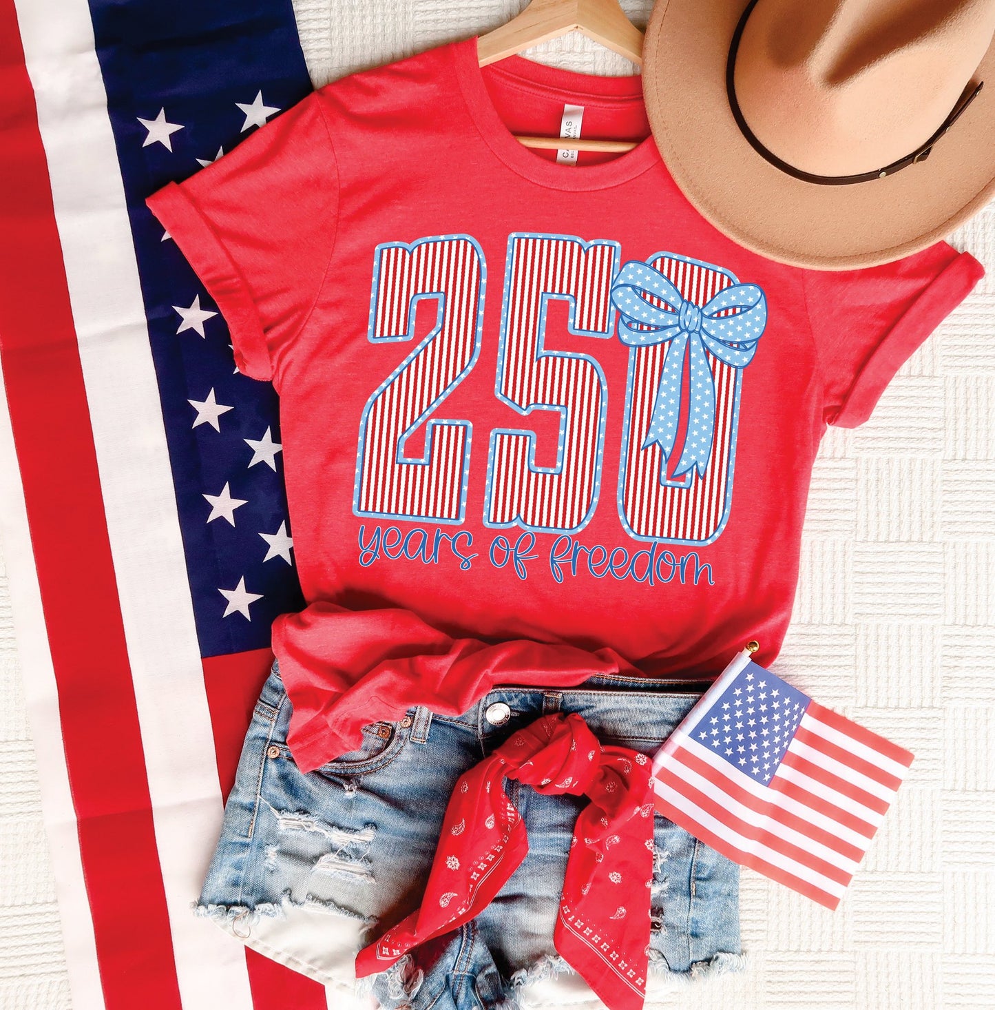 Red 250 Years of Freedom patriotic graphic tee styled with denim shorts, American flag, red bandana, and tan hat.