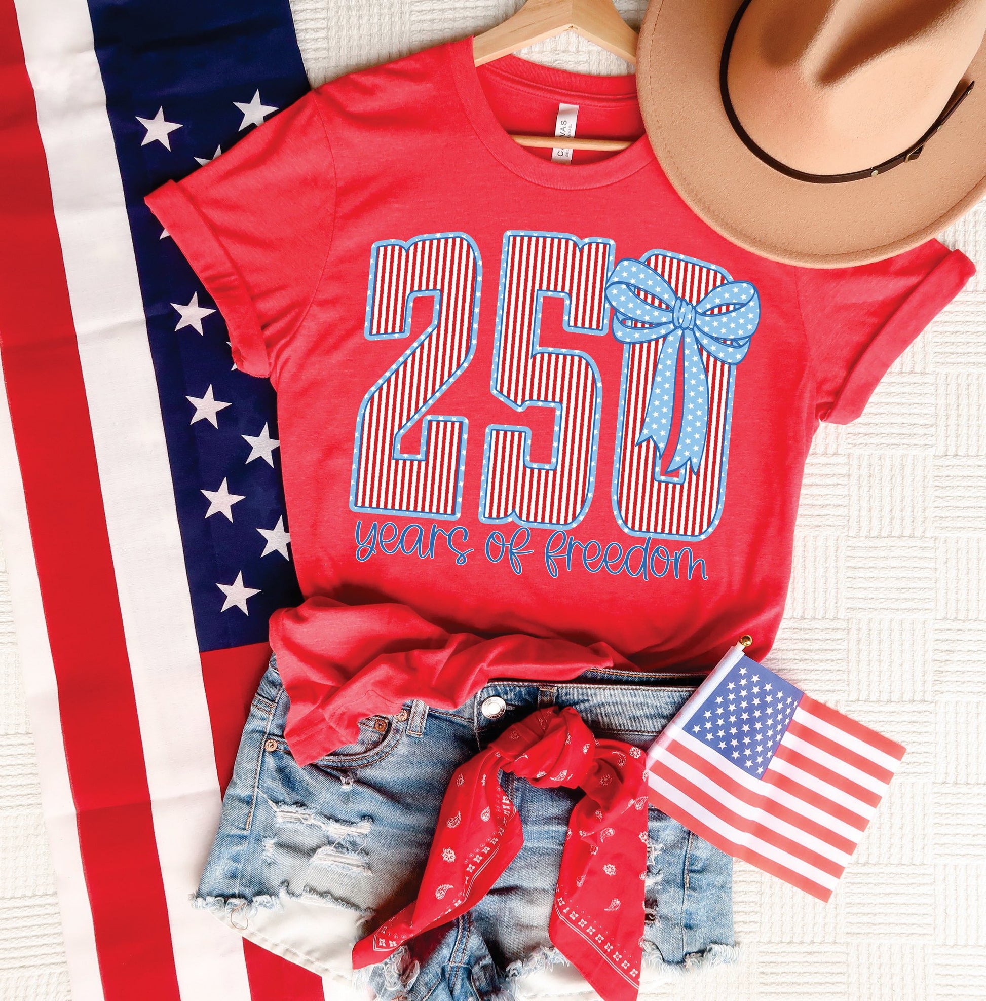 Red 250 Years of Freedom patriotic graphic tee styled with denim shorts, American flag, red bandana, and tan hat.