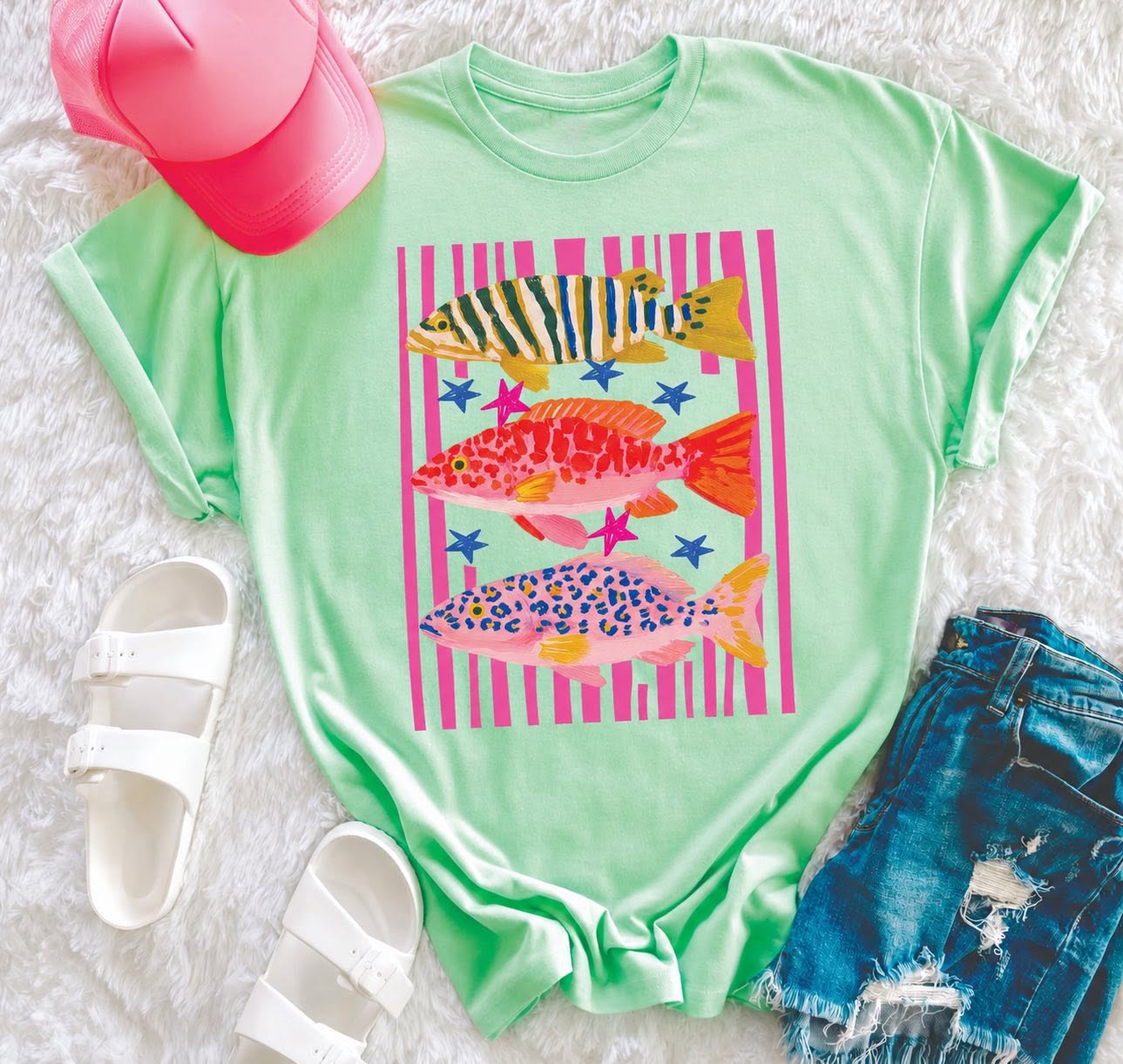 Mint green summer graphic tee with colorful preppy fish design, pink stripes, and star accents, styled with denim shorts, white sandals, and a pink hat.