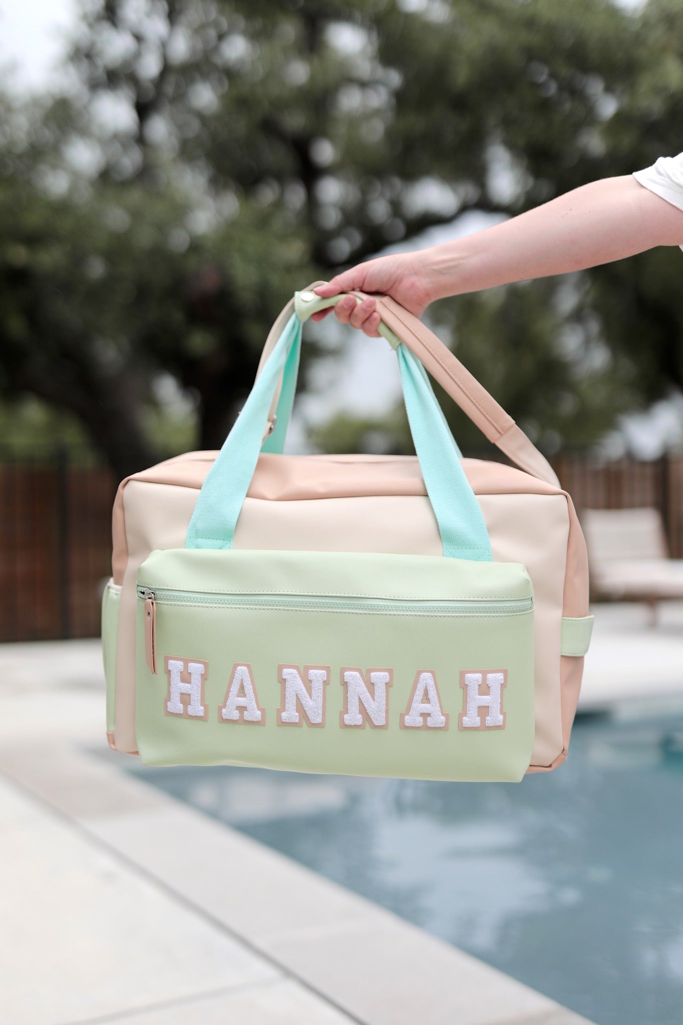Light pink and green duffel bag with 'HANNAH' printed on it, held by a hand by a poolside.