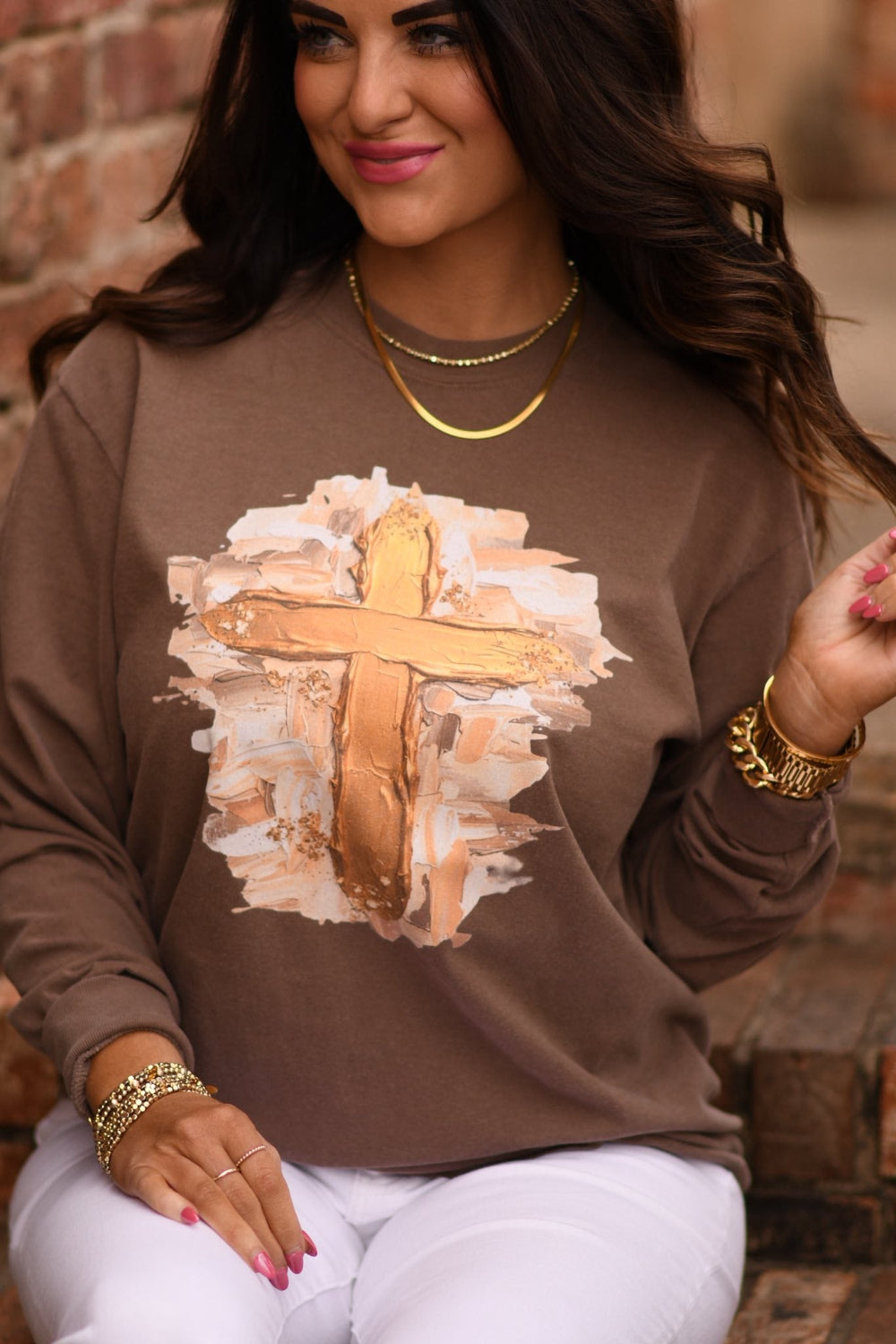 Woman wearing an espresso-colored Comfort Colors sweatshirt with a gold smudge cross design, styled with white pants and gold jewelry, sitting on brick steps.