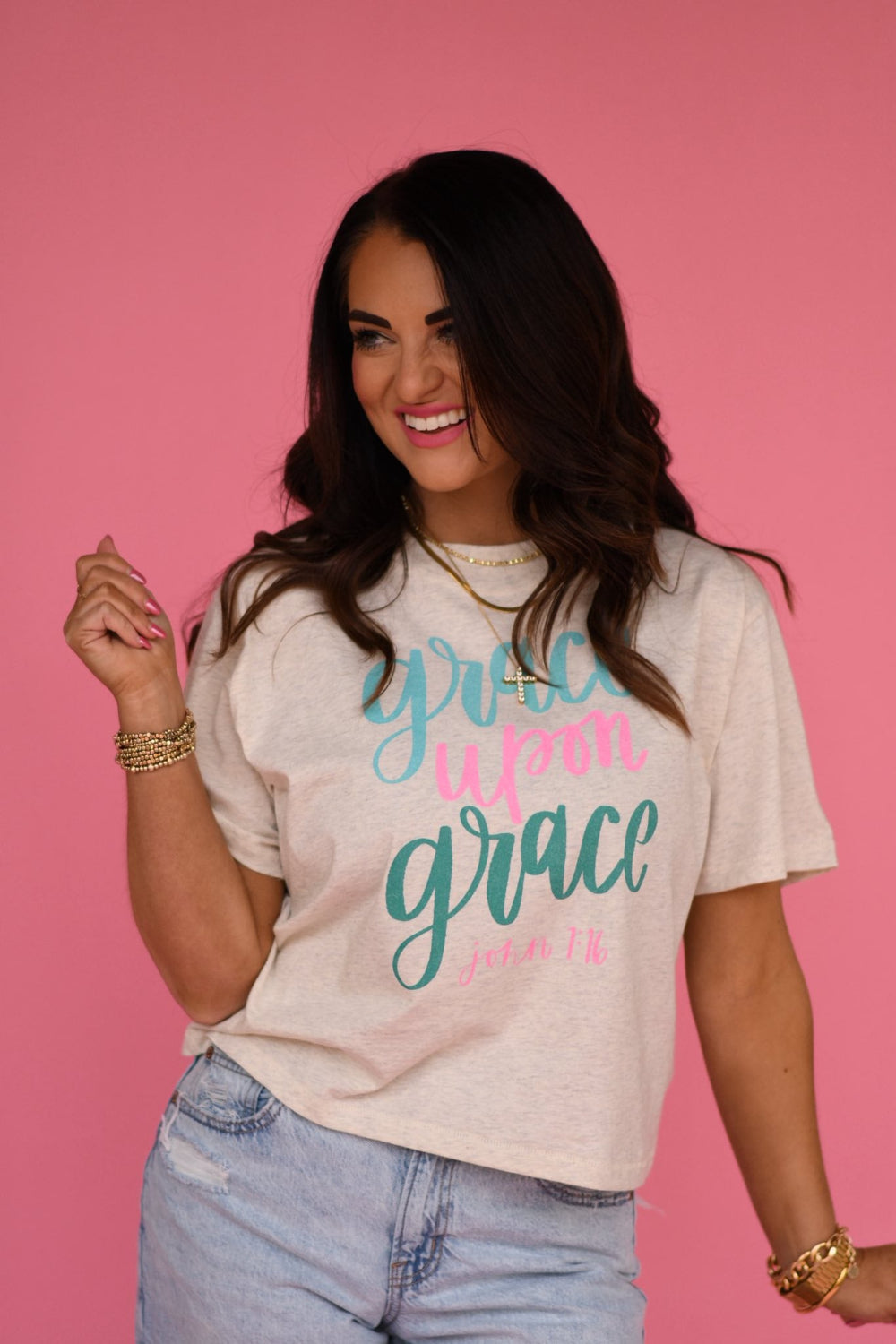 Woman wearing a cream-colored “Grace Upon Grace” Christian graphic tee with teal and pink lettering, styled with denim jeans against a pink background.