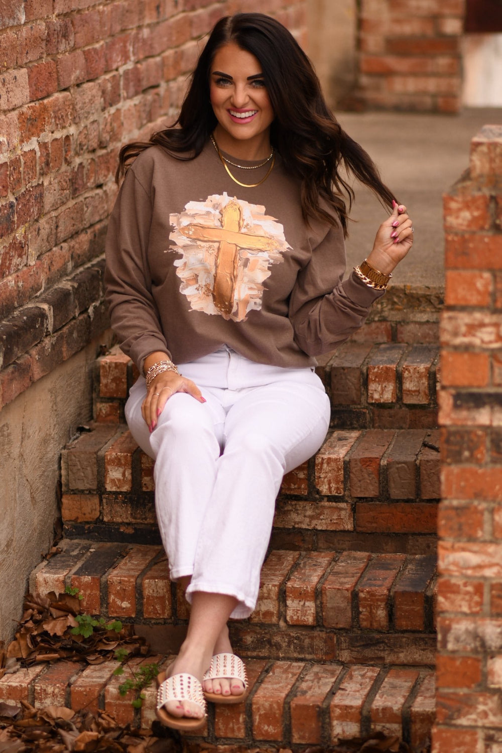 Woman wearing an espresso-colored Comfort Colors sweatshirt with a gold smudge cross design, styled with white pants and gold jewelry, sitting on brick steps.