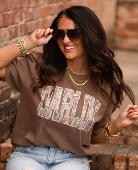 Woman wearing a brown Comfort Colors unisex graphic tee with camo “Darlin’” lettering styled with denim jeans and gold accessories