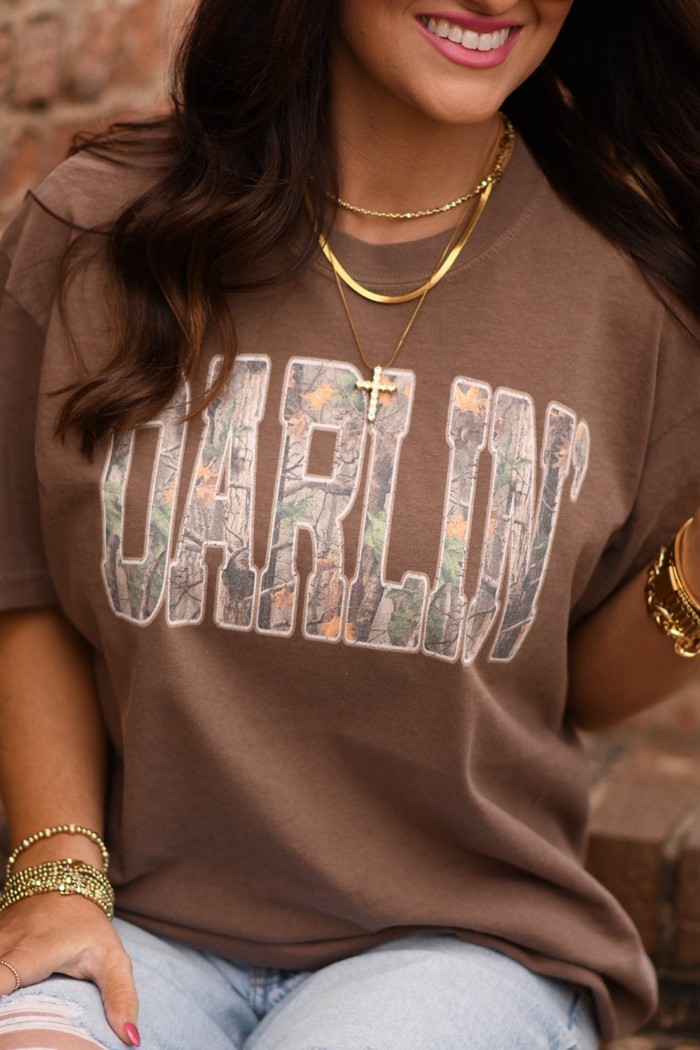 Woman wearing a brown Comfort Colors unisex graphic tee with camo “Darlin’” lettering styled with denim jeans and gold accessories