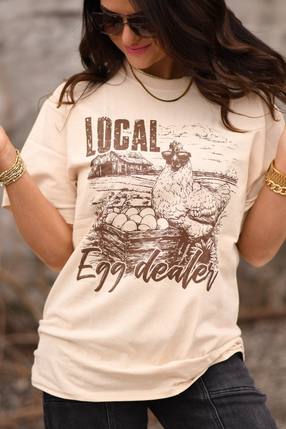 Tan graphic t-shirt with a chicken illustration and the phrase “Local Egg Dealer,” worn by a woman, featuring a rustic farm-style design.