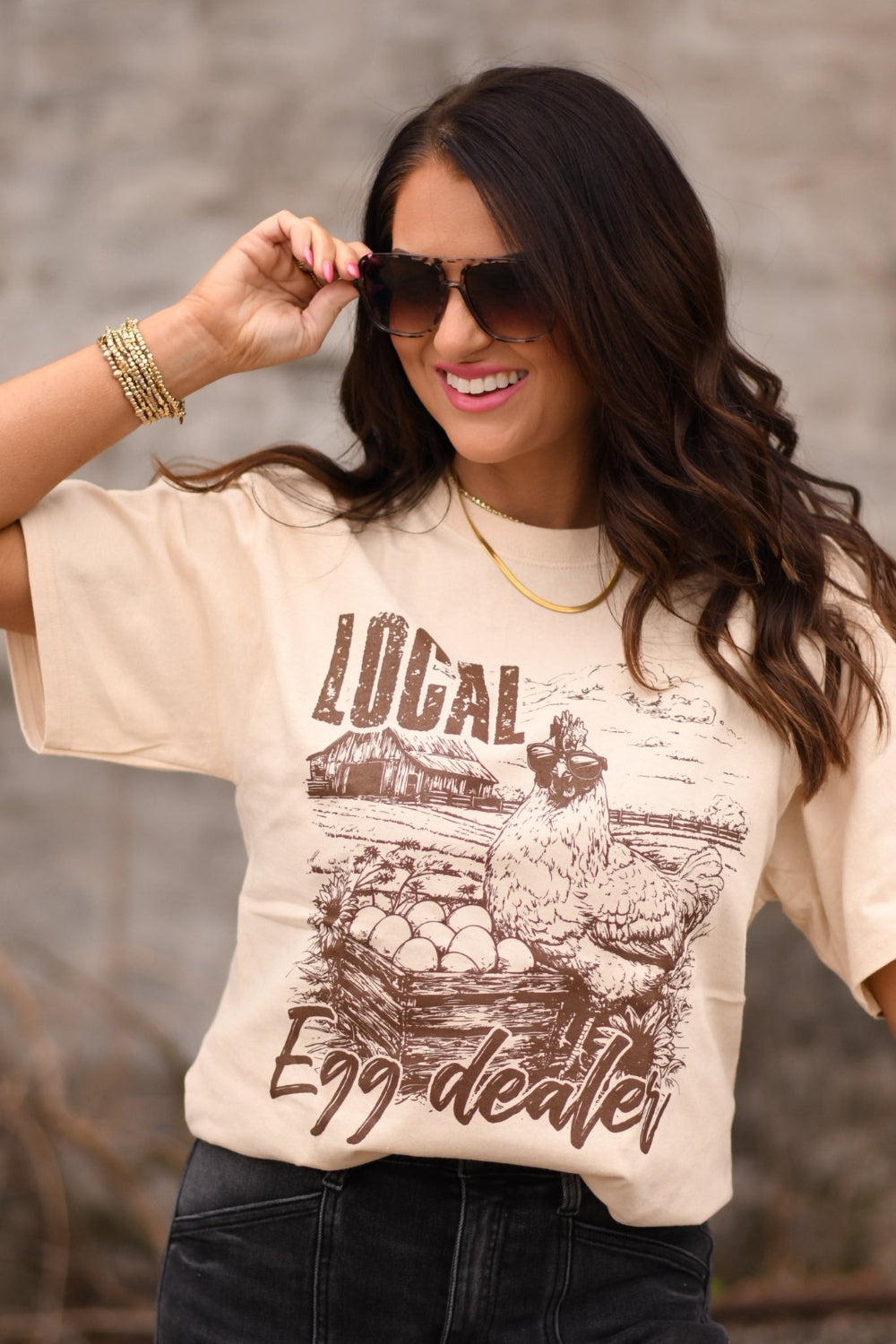 Tan graphic t-shirt with a chicken illustration and the phrase “Local Egg Dealer,” worn by a woman, featuring a rustic farm-style design.