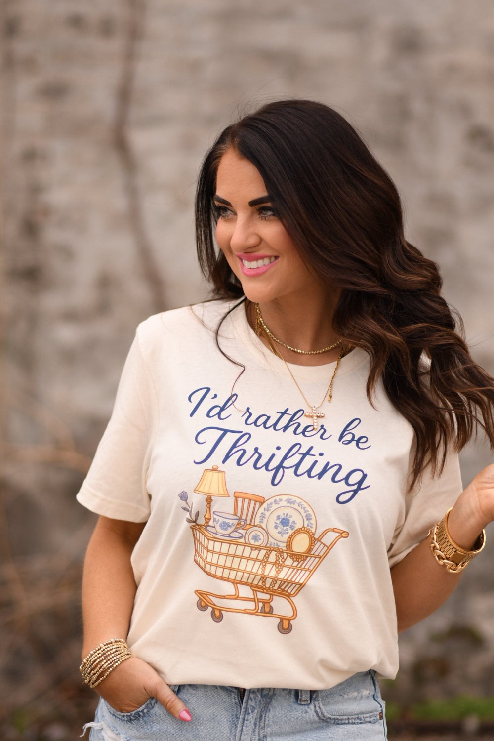 Woman wearing a natural colored graphic t-shirt that says “I’d Rather Be Thrifting” with a vintage shopping cart illustration, styled with jeans and gold jewelry