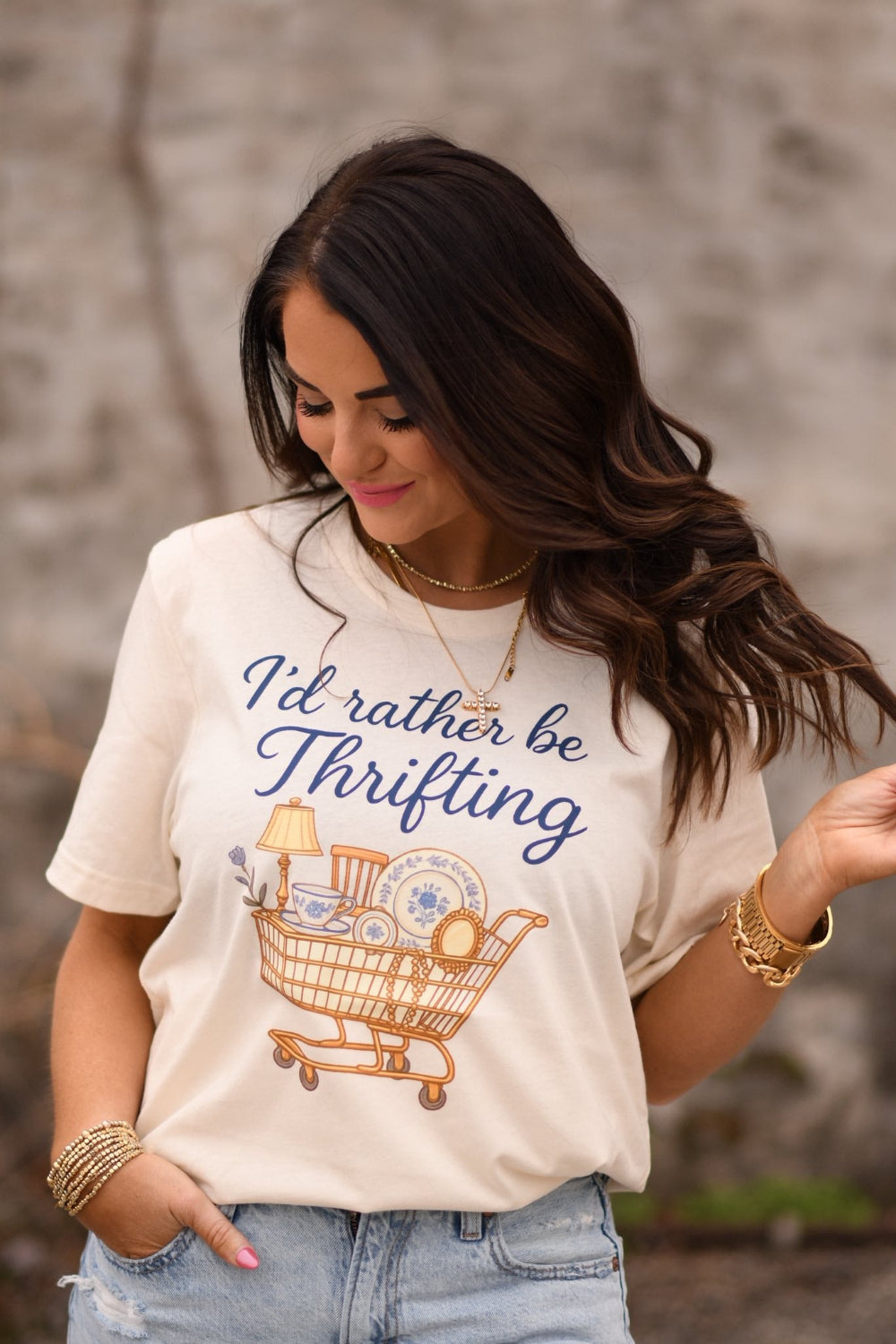 Woman wearing a natural colored graphic t-shirt that says “I’d Rather Be Thrifting” with a vintage shopping cart illustration, styled with jeans and gold jewelry