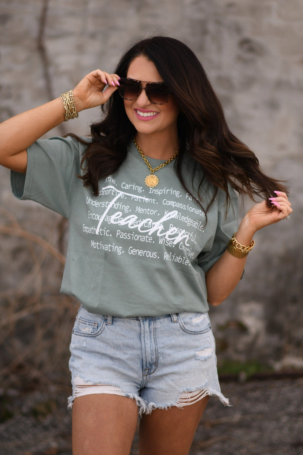 Woman modeling sage Teacher Delta Tee with inspirational teacher words, unisex Delta Pro graphic shirt, styled with distressed denim shorts and gold accessories