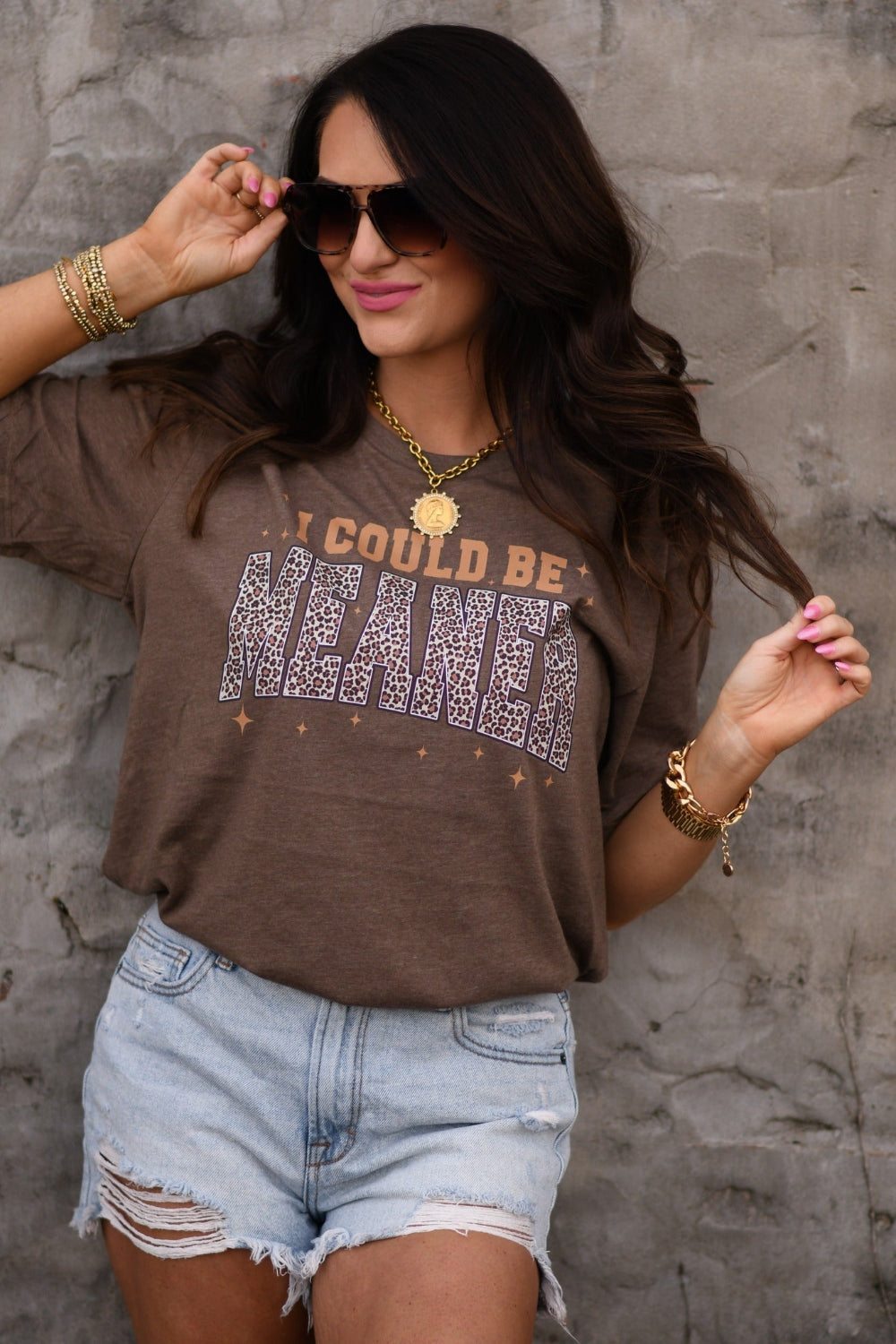 Woman wearing heather brown I Could Be Meaner leopard graphic tee on Bella Canvas unisex t-shirt with distressed denim shorts and gold jewelry