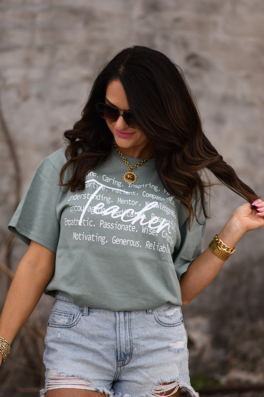 Woman modeling sage Teacher Delta Tee with inspirational teacher words, unisex Delta Pro graphic shirt, styled with distressed denim shorts and gold accessories