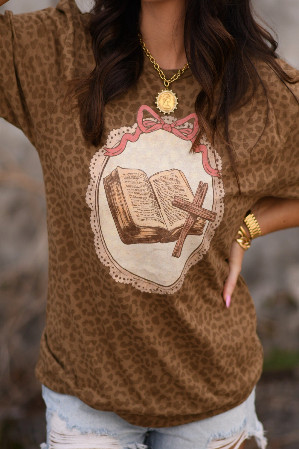 Woman wearing a leopard print Christian graphic tee featuring an open Bible and cross design, styled with light wash distressed jeans and gold jewelry.