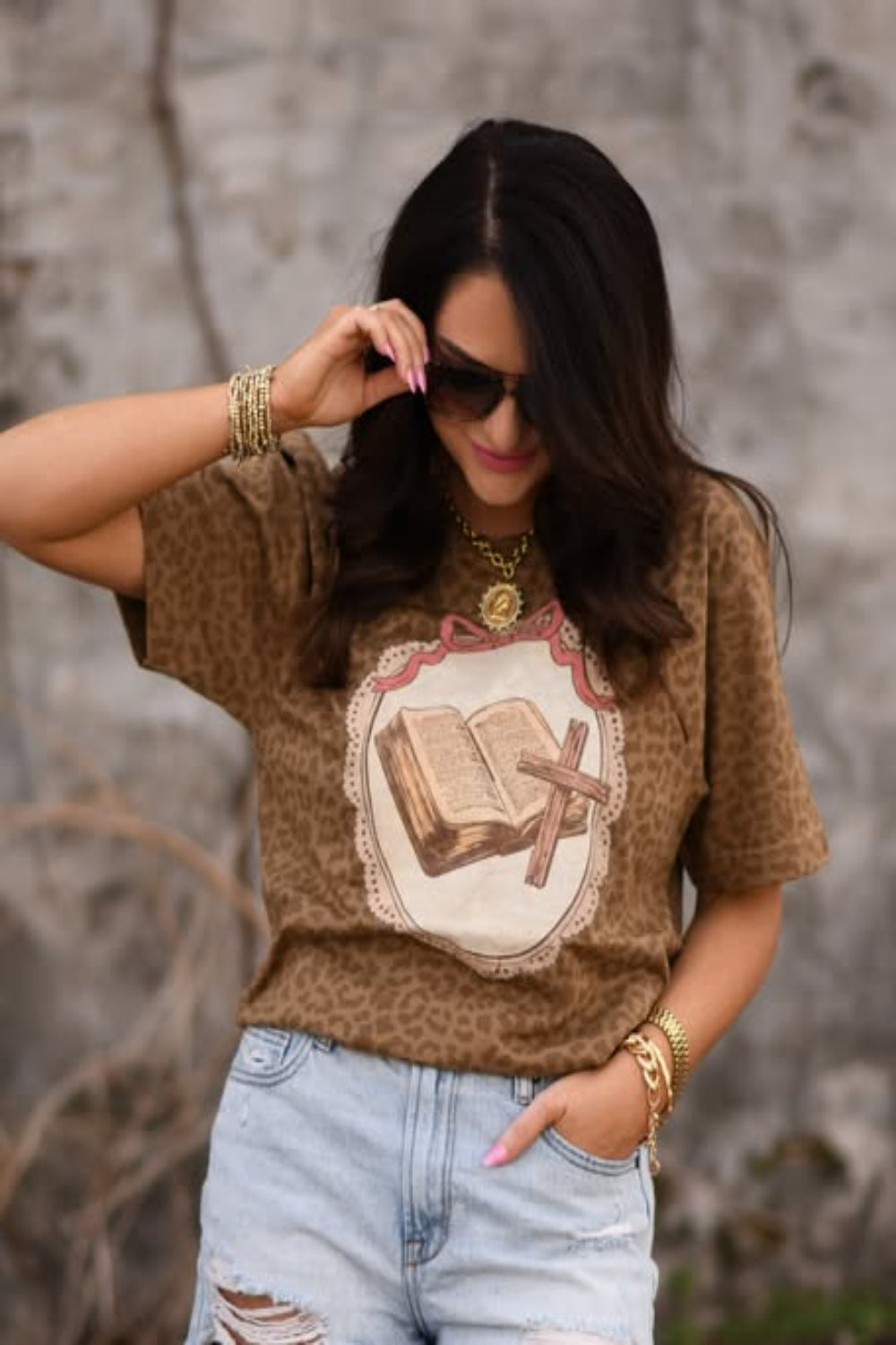 Woman wearing a leopard print Christian graphic tee featuring an open Bible and cross design, styled with light wash distressed jeans and gold jewelry.