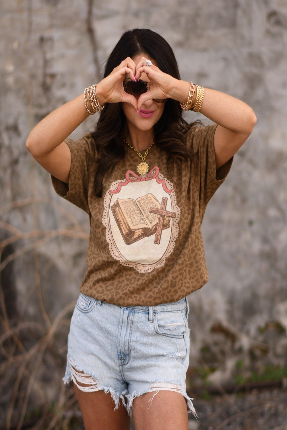 Woman wearing a leopard print Christian graphic tee featuring an open Bible and cross design, styled with light wash distressed jeans and gold jewelry.
