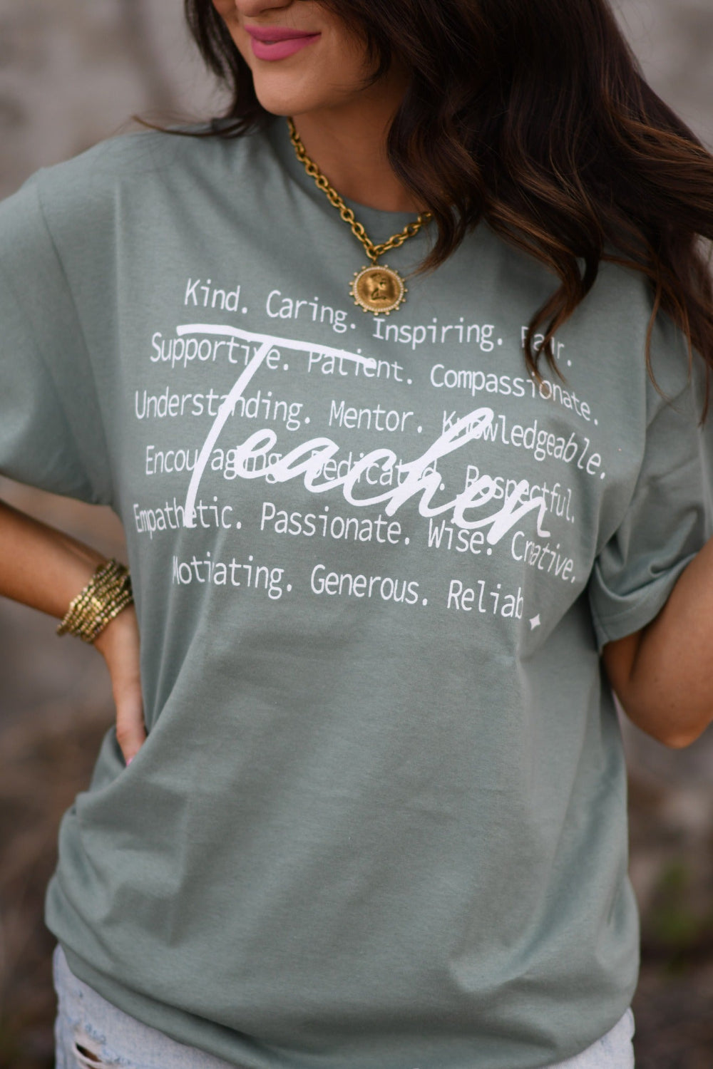 Woman modeling sage Teacher Delta Tee with inspirational teacher words, unisex Delta Pro graphic shirt, styled with distressed denim shorts and gold accessories