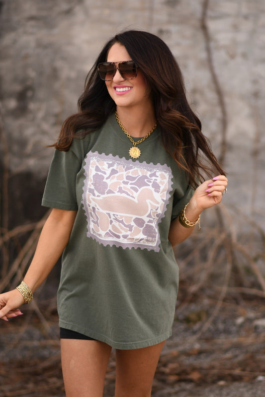 Woman wearing moss green Comfort Colors unisex t-shirt featuring duck camo stamp graphic, styled with sunglasses and gold jewelry.