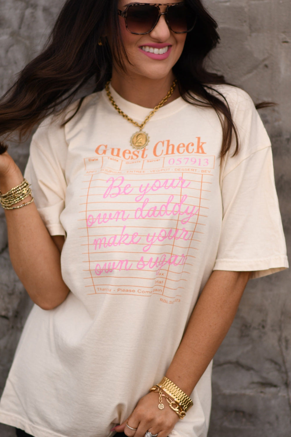 Woman modeling ivory Comfort Colors graphic tee with pink “Be Your Own Daddy Make Your Own Sugar” design, styled with sunglasses and gold jewelry.