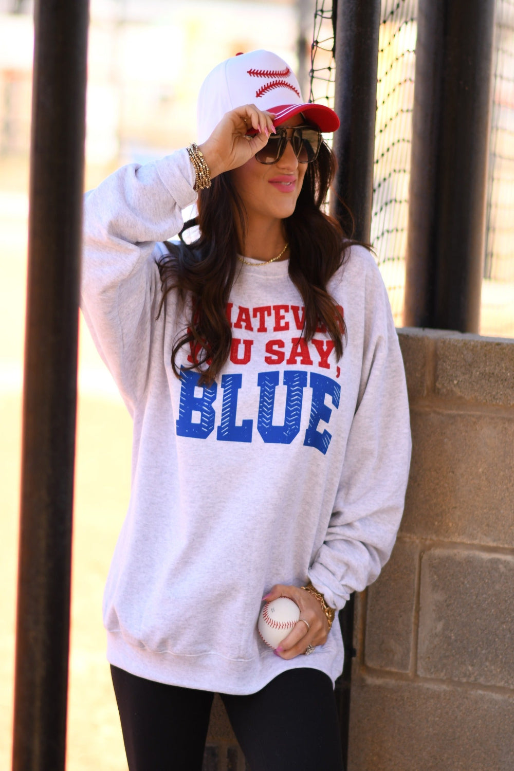 Woman wearing ash gray “Whatever You Say Blue” baseball graphic sweatshirt with red and blue lettering, styled with a baseball cap and holding a baseball at a ball field.
