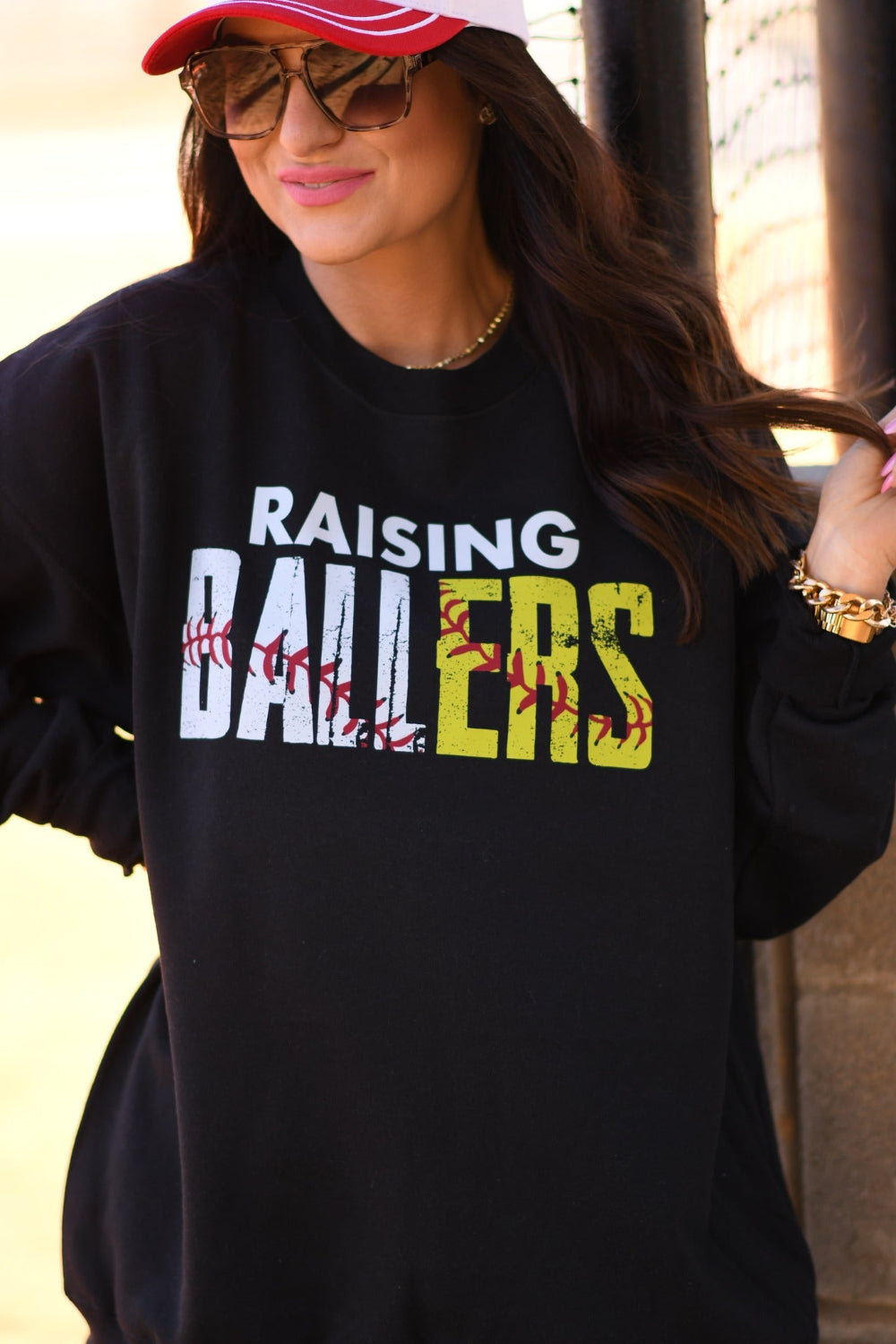 Woman wearing black Raising Ballers baseball mom sweatshirt with distressed white and yellow lettering and baseball stitch graphic, styled with red cap and sunglasses.