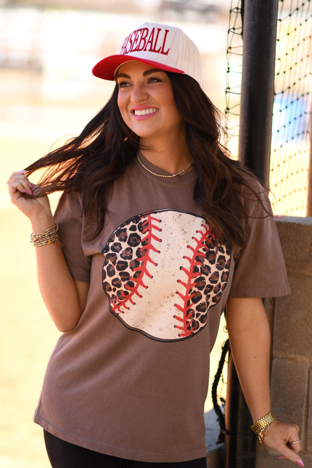 Woman wearing an espresso Comfort Colors leopard baseball graphic tee with red stitching and a white baseball cap at a baseball field.