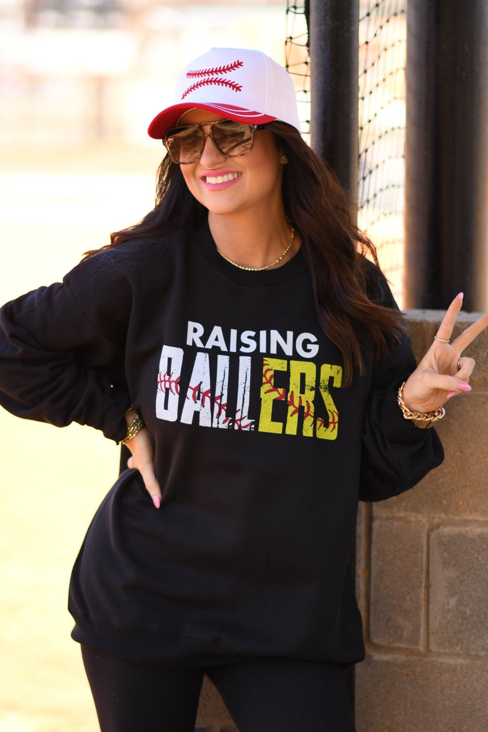 Woman wearing black Raising Ballers baseball and softball graphic sweatshirt at ballpark with baseball hat