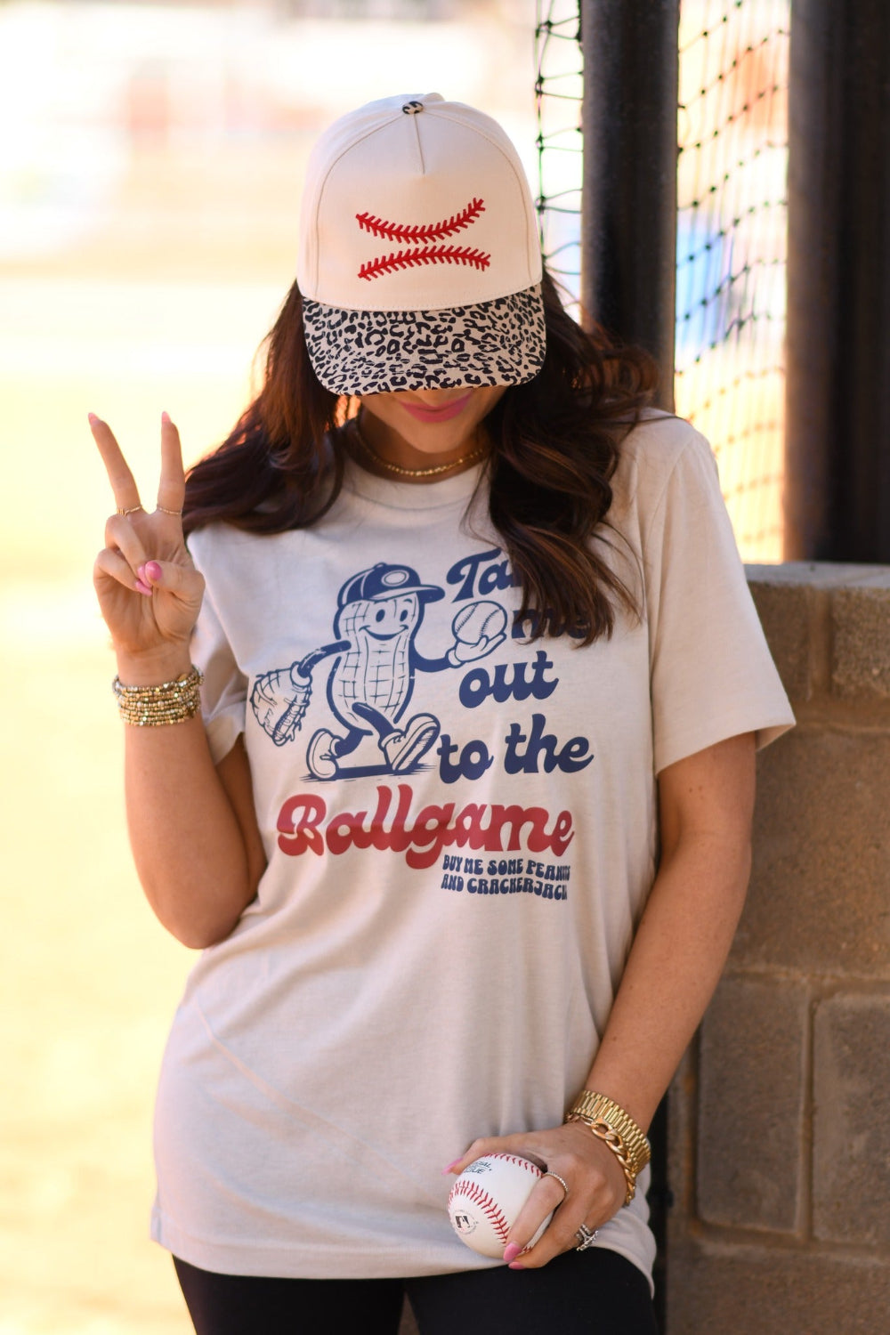 Woman wearing Heather Dust Bella Canvas “Take Me Out To The Ballgame” peanut baseball graphic tee with leopard brim baseball hat at a baseball field.