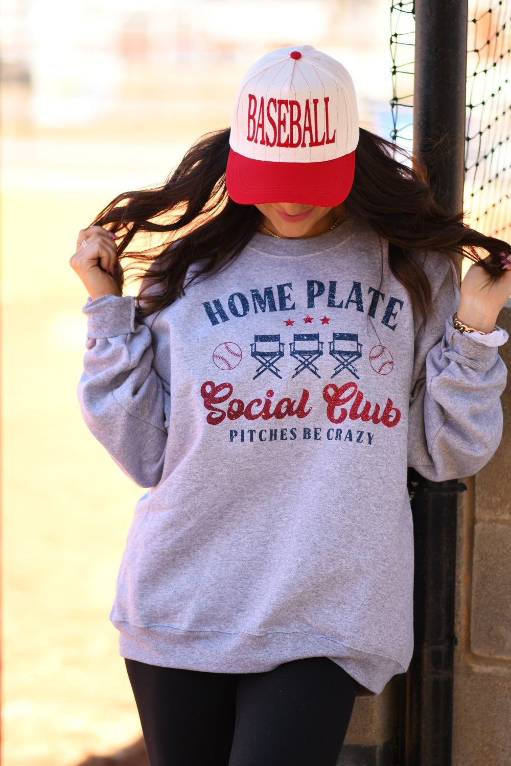 Woman wearing a Heather Athletic Grey Home Plate Social Club baseball graphic sweatshirt with red and navy print and a baseball cap at a baseball field