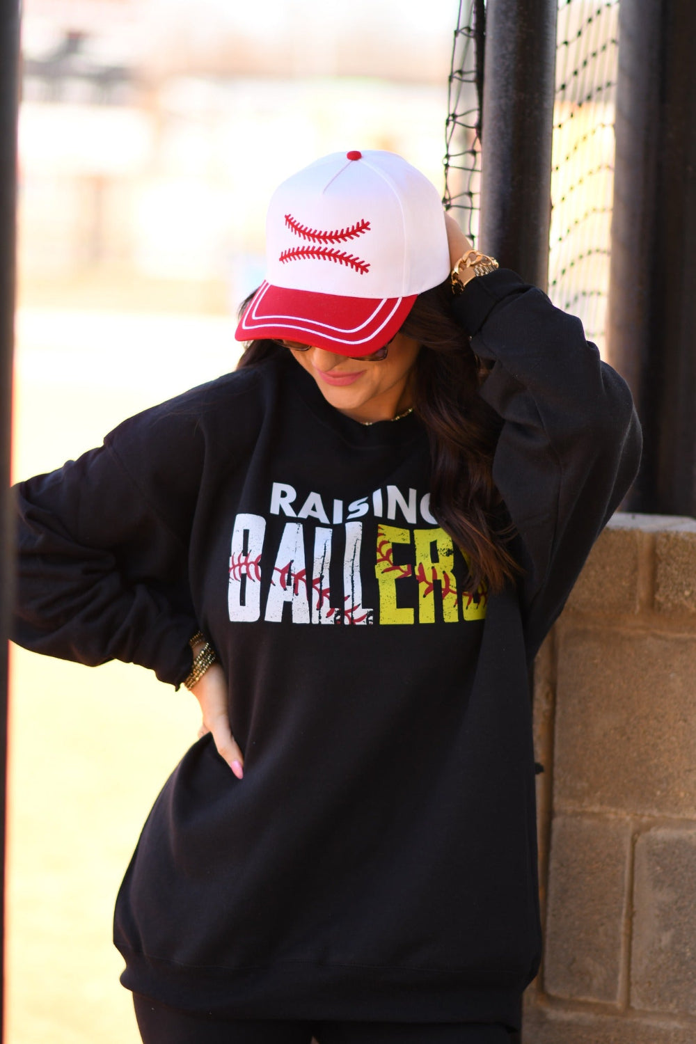Woman wearing black Raising Ballers baseball and softball graphic sweatshirt at ballpark with baseball hat