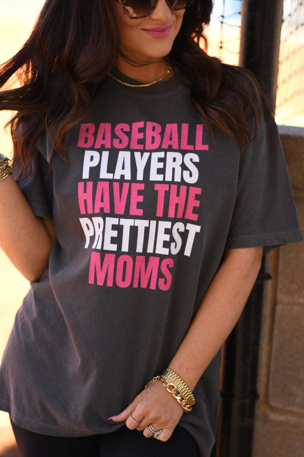 Woman wearing a pepper Comfort Colors graphic tee that says "Baseball Players Have The Prettiest Moms" in pink and white lettering, styled with sunglasses and black leggings at a baseball field.
