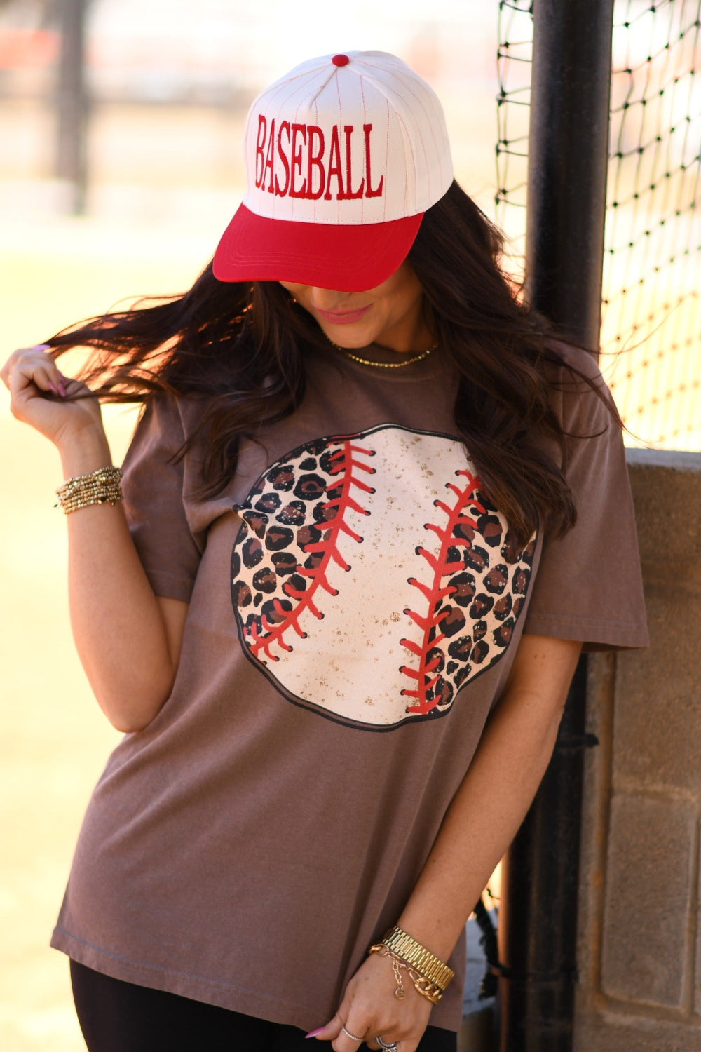 Woman wearing an espresso Comfort Colors leopard baseball graphic tee with red stitching and a white baseball cap at a baseball field.