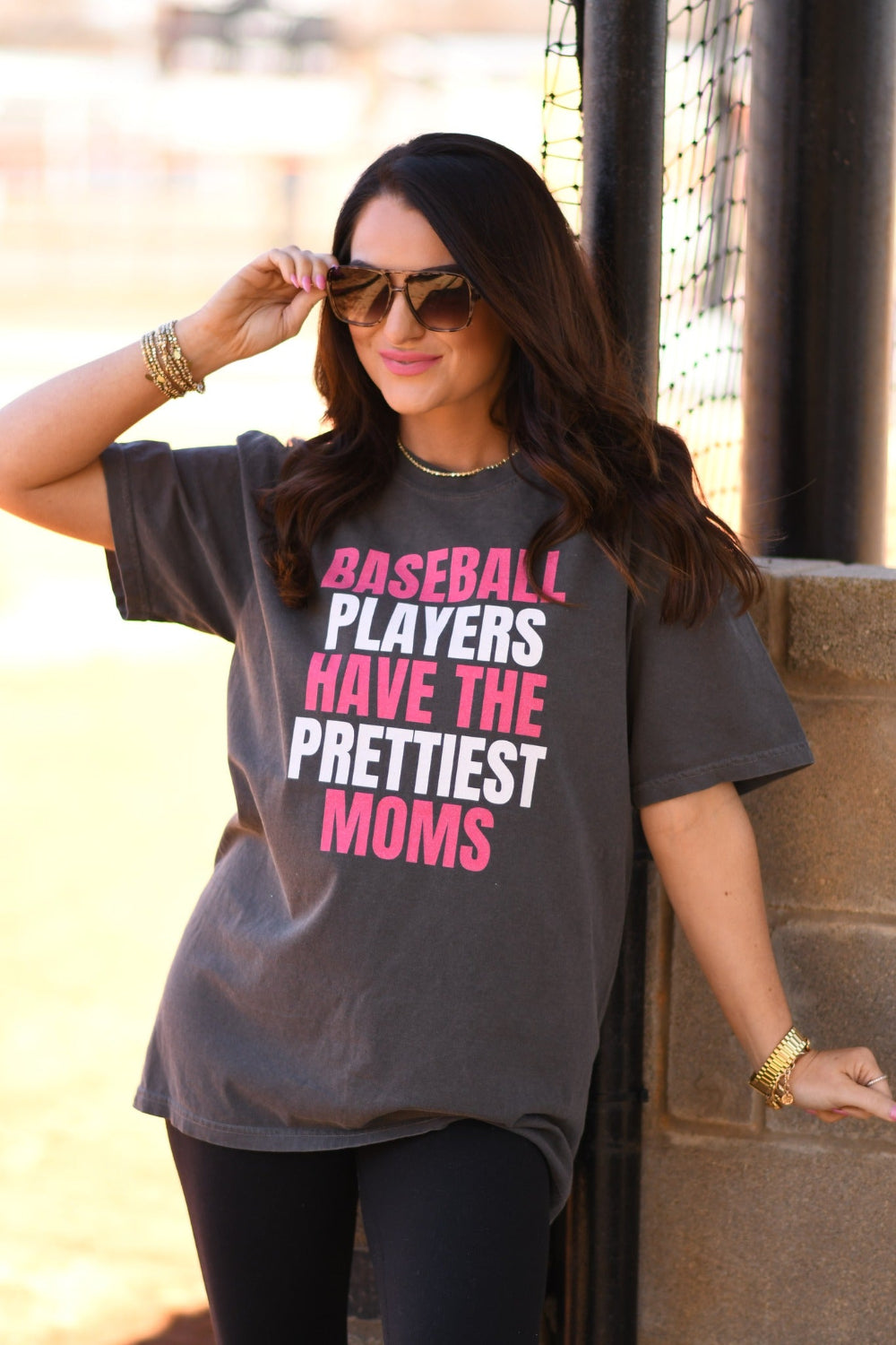 Woman wearing a pepper Comfort Colors graphic tee that says "Baseball Players Have The Prettiest Moms" in pink and white lettering, styled with sunglasses and black leggings at a baseball field.
