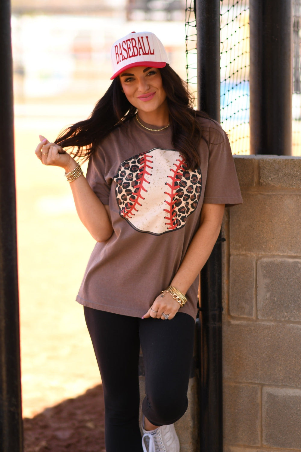 Woman wearing an espresso Comfort Colors leopard baseball graphic tee with red stitching and a white baseball cap at a baseball field.