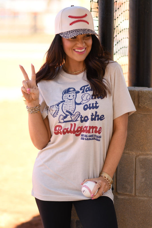 Woman wearing Heather Dust Bella Canvas “Take Me Out To The Ballgame” peanut baseball graphic tee with leopard brim baseball hat at a baseball field.