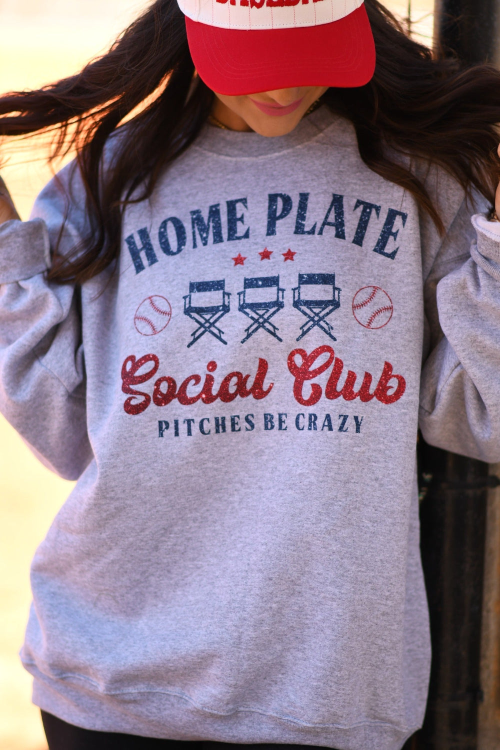 Woman wearing a Heather Athletic Grey Home Plate Social Club baseball graphic sweatshirt with red and navy print and a baseball cap at a baseball field
