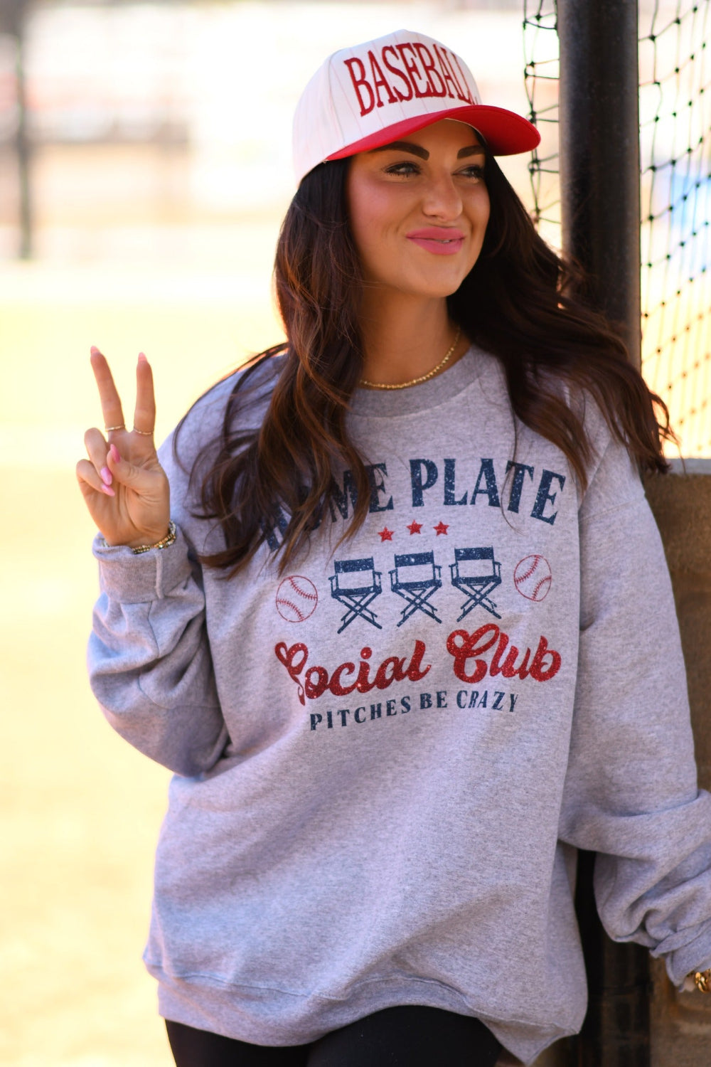 Woman wearing a Heather Athletic Grey Home Plate Social Club baseball graphic sweatshirt with red and navy print and a baseball cap at a baseball field