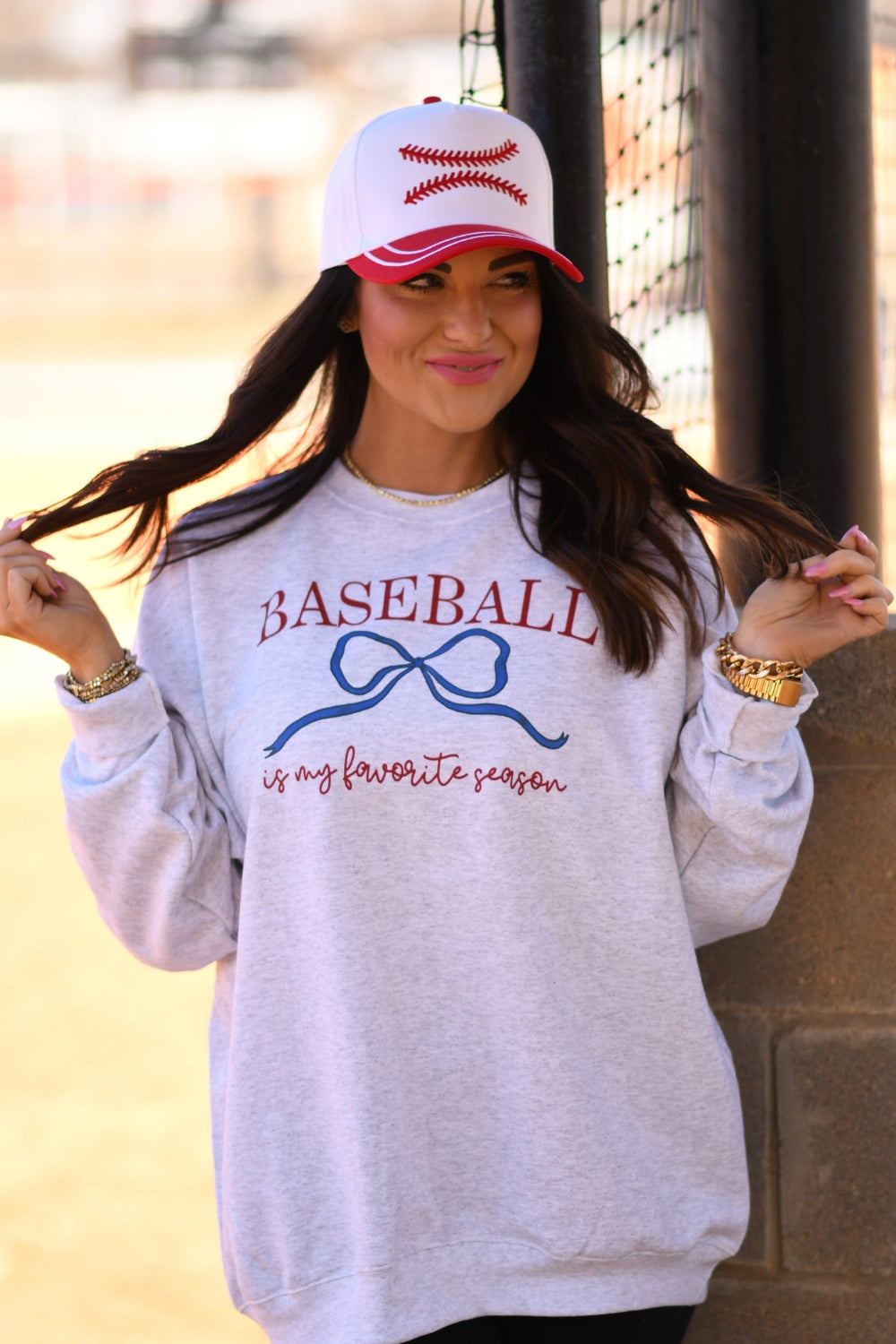 Woman wearing ash gray “Baseball Is My Favorite Season” graphic sweatshirt with blue bow design and red baseball hat at baseball field.