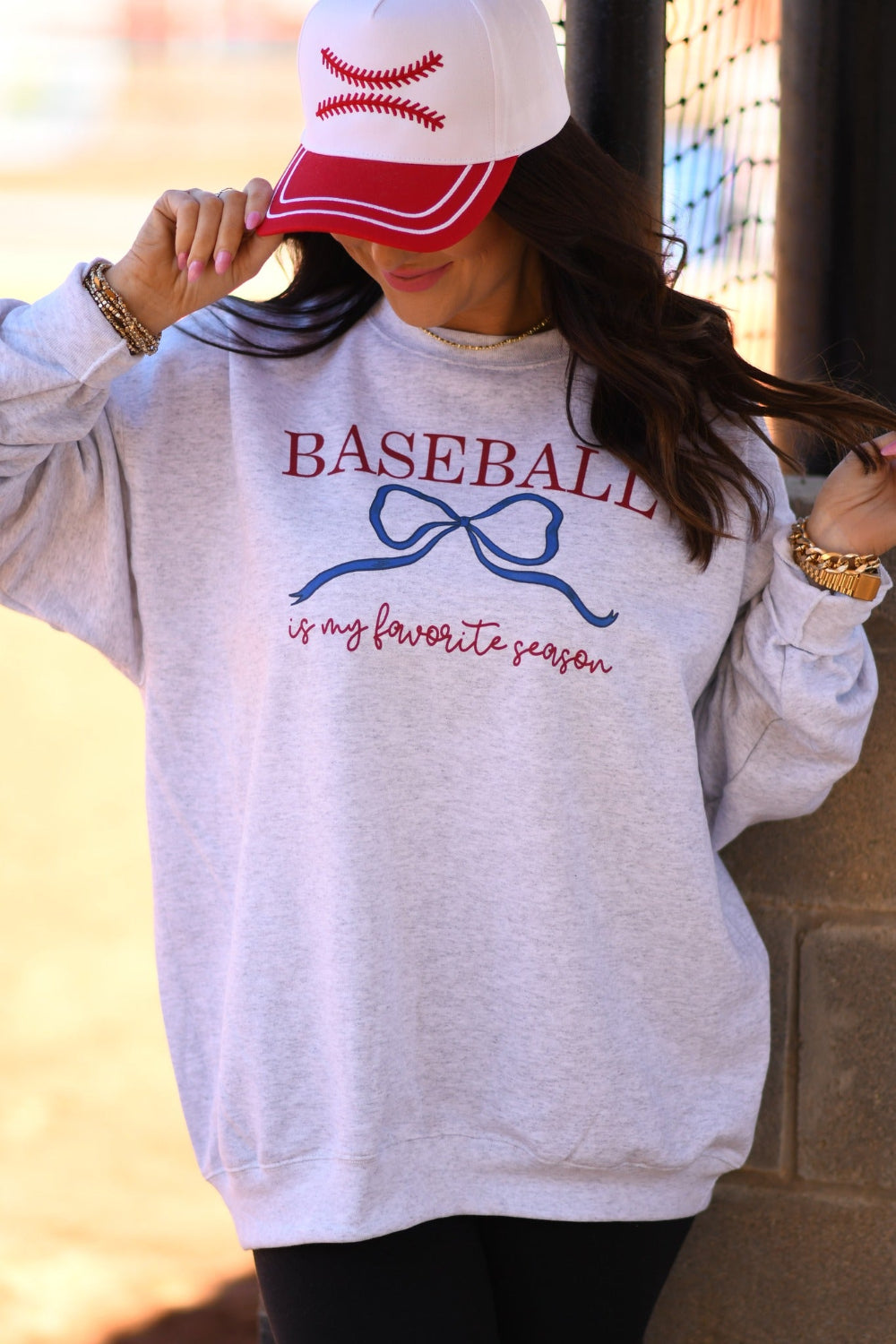 Woman wearing ash gray “Baseball Is My Favorite Season” graphic sweatshirt with blue bow design and red baseball hat at baseball field.